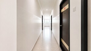 Un pasillo largo y estrecho con paredes blancas y suelo de baldosas, que lleva a una puerta con un letrero rojo.