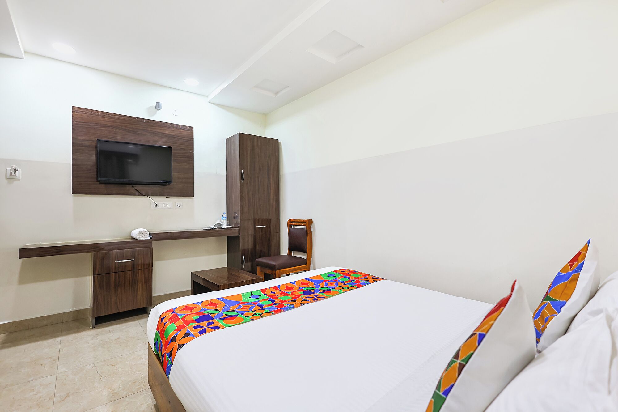 A hotel room with a bed, a desk with a chair, a television mounted on the wall, and a wardrobe.