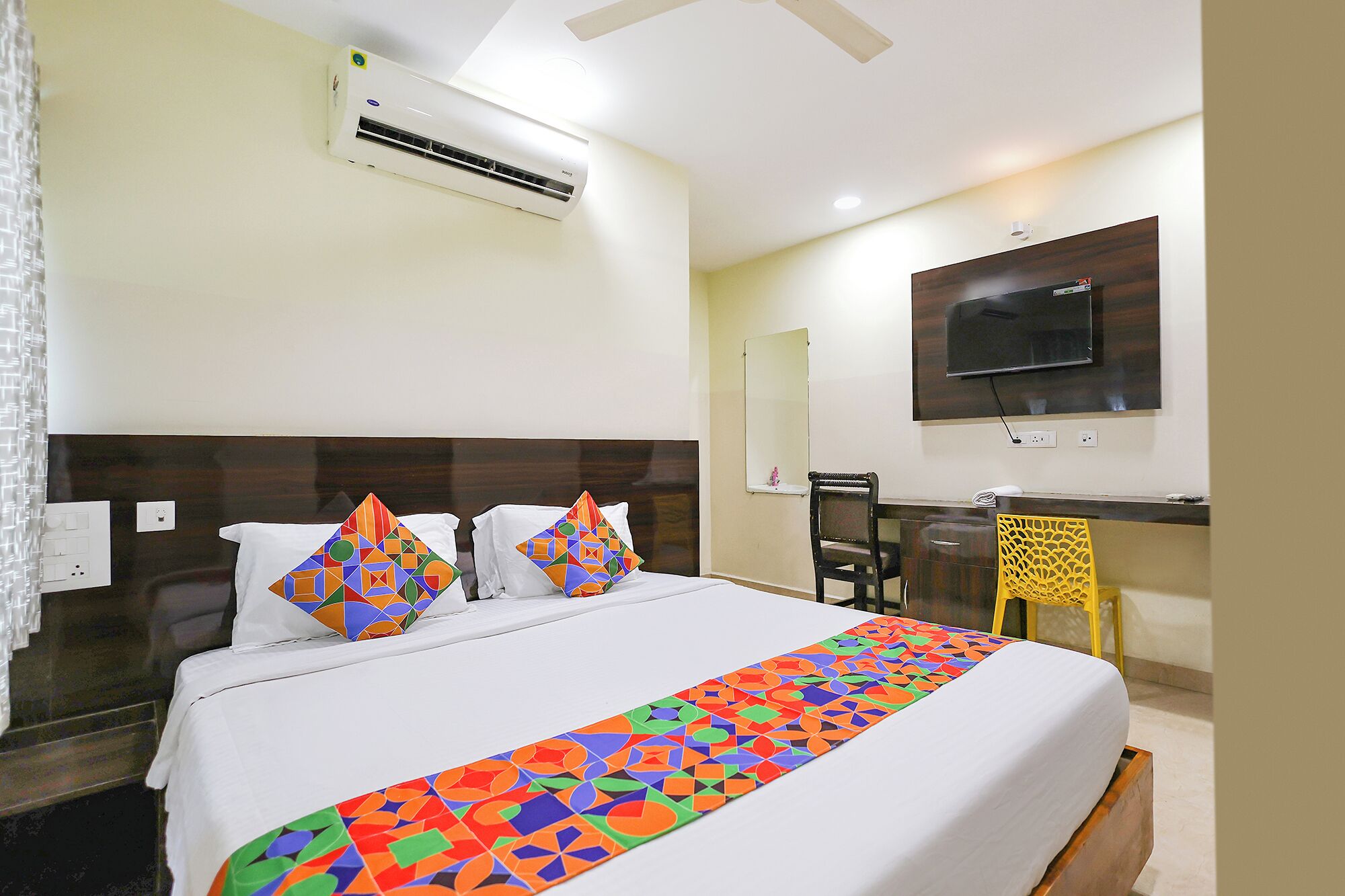 A hotel room with a bed, an air conditioner, a desk with a chair, a TV, and a mirror.