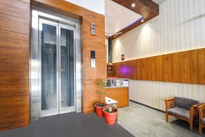 A reception area with a wooden reception desk, a seating area with a sofa and chairs, a glass elevator, and a wall-mounted sign. - Fabhotel Amar Residency (New Delhi)