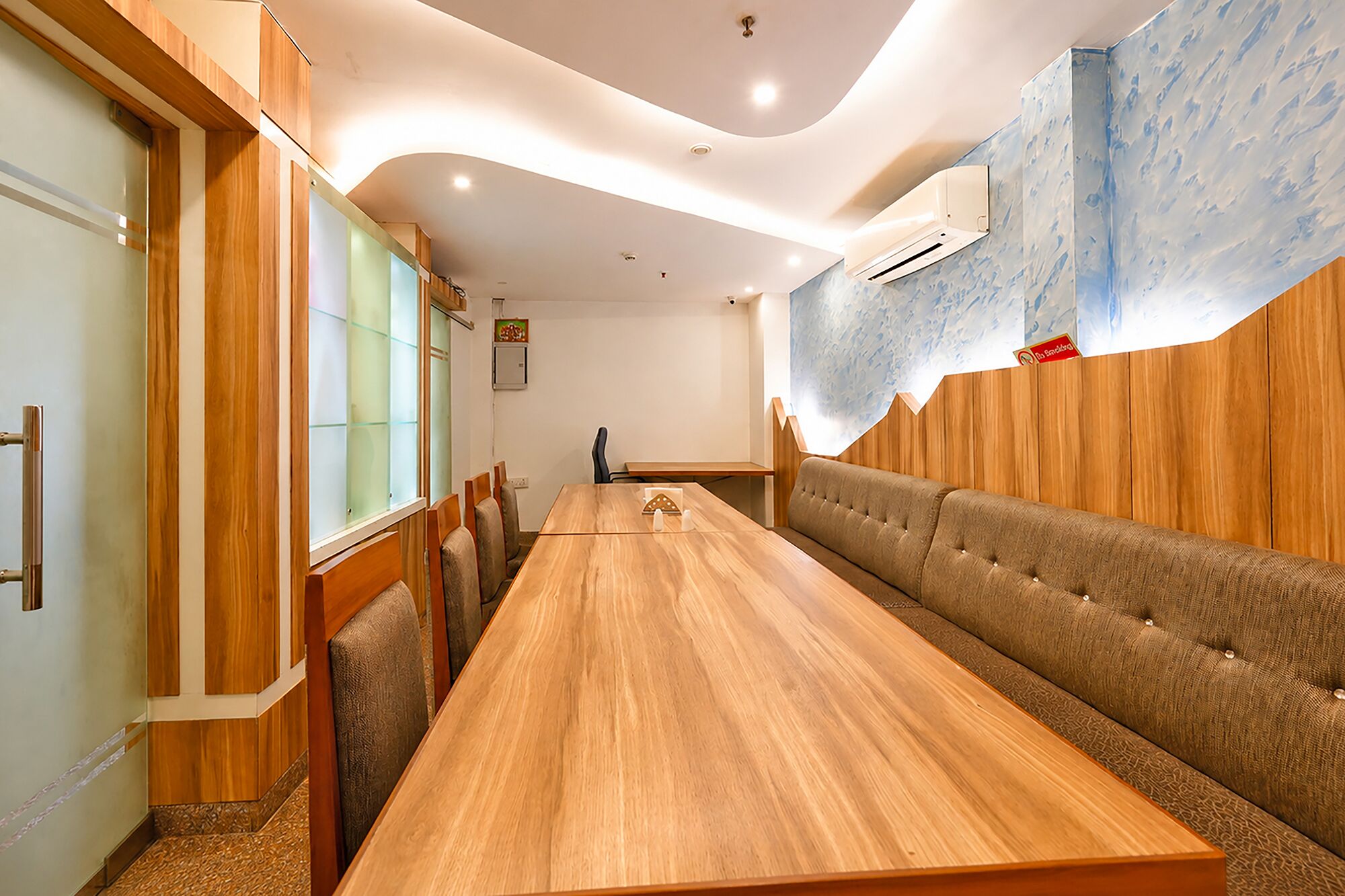 A long wooden table with cushioned seating on both sides, a ceiling with recessed lighting, and a wall-mounted air conditioning unit.