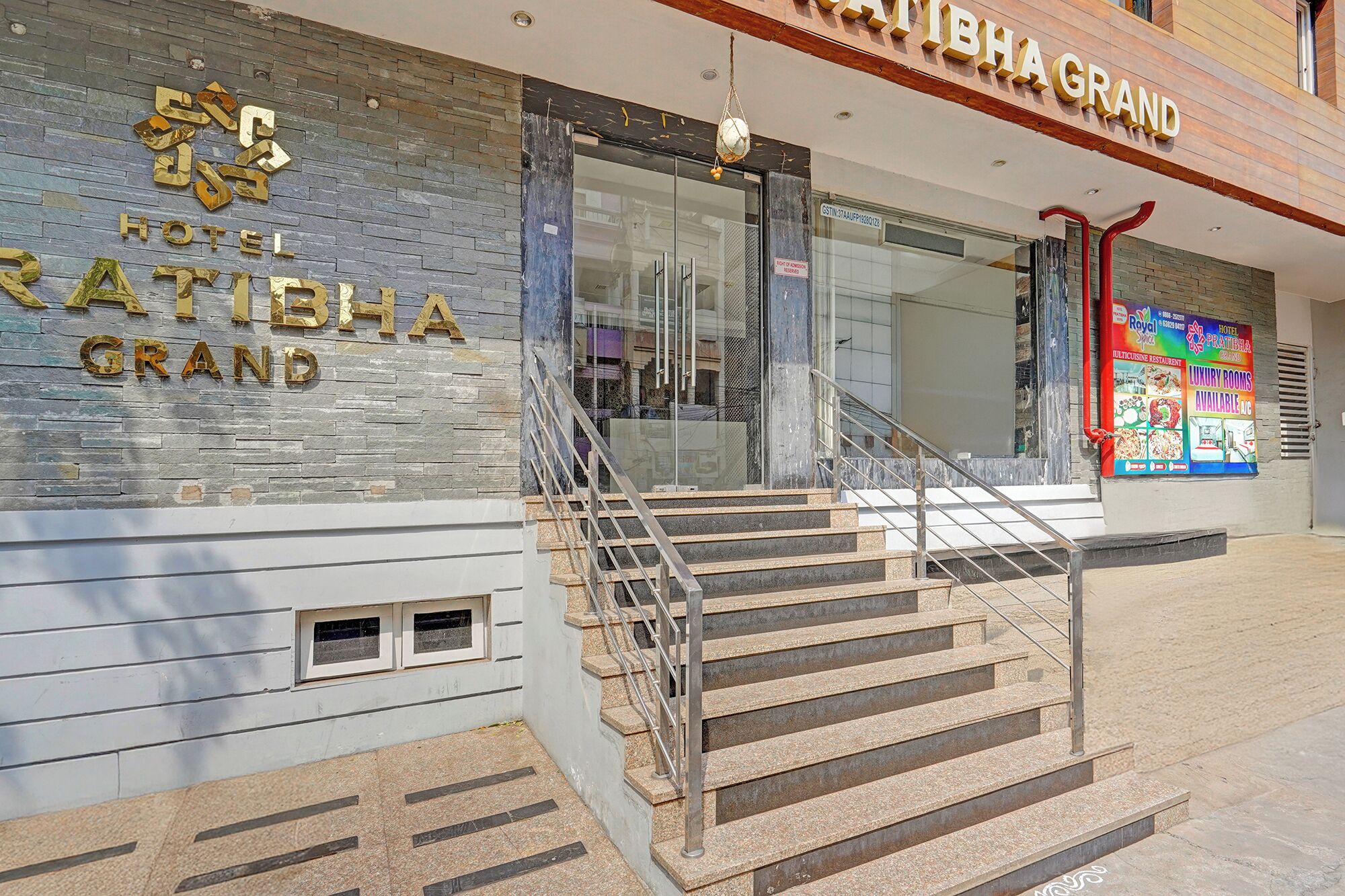 The exterior of Hotel Ratibha Grand with a stone facade, a staircase leading to the entrance, and a signboard advertising the hotel and its amenities.
