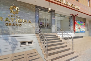 Exterior del Hotel Ratibha Grand, con fachada de piedra, una escalera que conduce a la entrada y un cartel que anuncia el hotel y sus servicios.