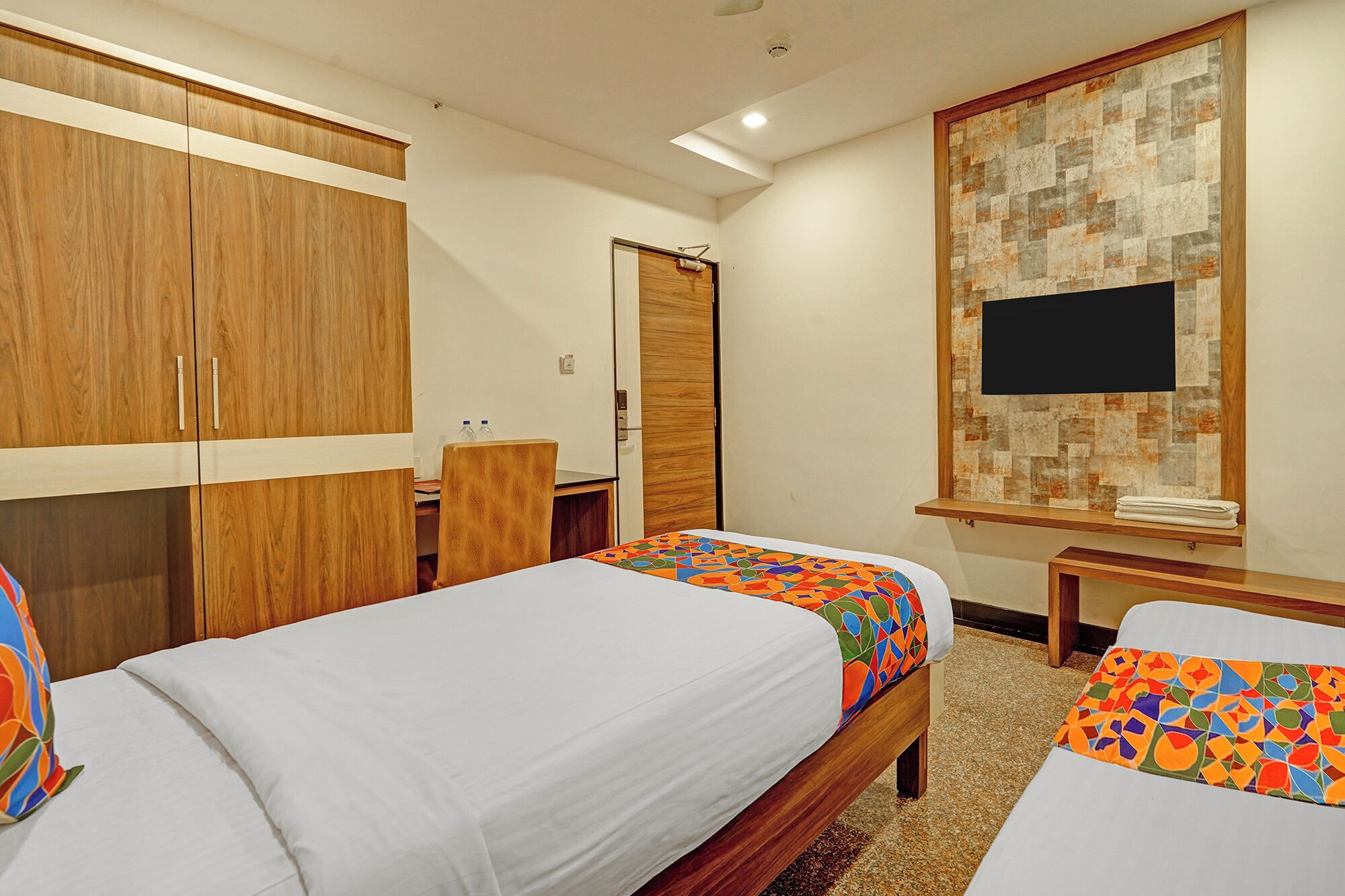 Une chambre d’hôtel avec deux lits, une armoire, un bureau, une télévision et une étagère fixée au mur.