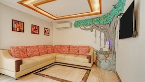 Uma sala de estar com um sofá seccional bege, teto de madeira com iluminação embutida, televisão fixada na parede e um mural decorativo com tema de árvore.