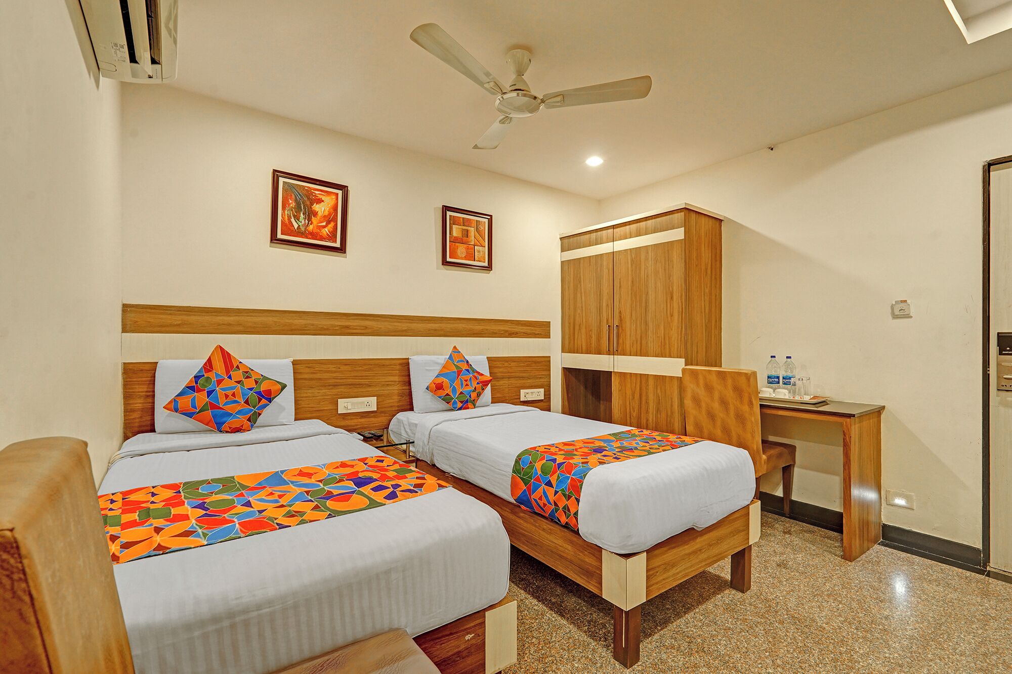 A hotel room with two beds, a wooden wardrobe, a desk, and a ceiling fan.