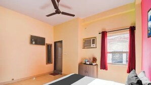 A bedroom with a bed, a ceiling fan, a window with curtains, a door, and a small table with a plant. - Fabhotel Radhy Stay (Jaipur)