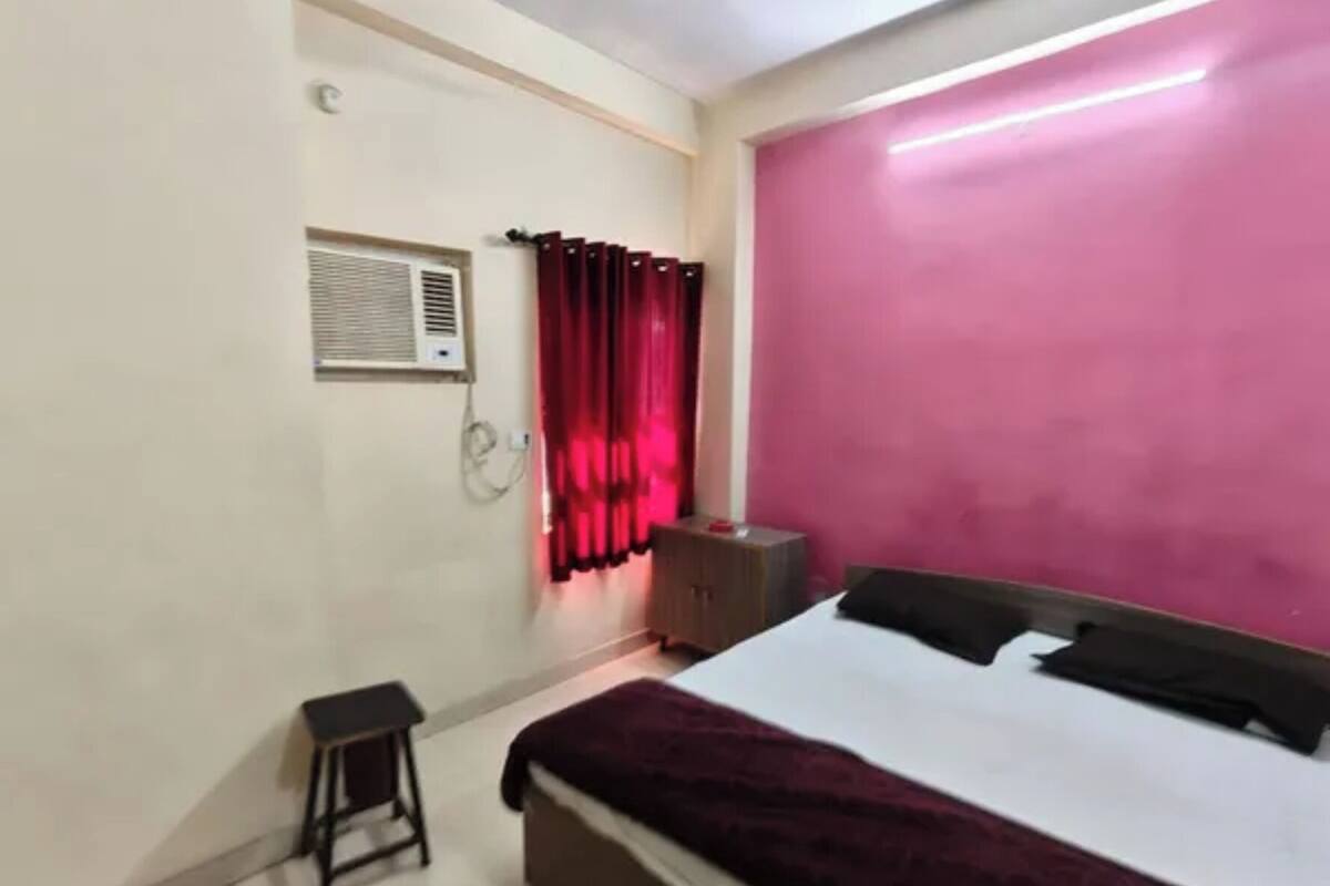 Ein Schlafzimmer mit Bett, Nachttisch, einem kleinen Hocker, einer Wand-Klimaanlage und einer rosa Wand.