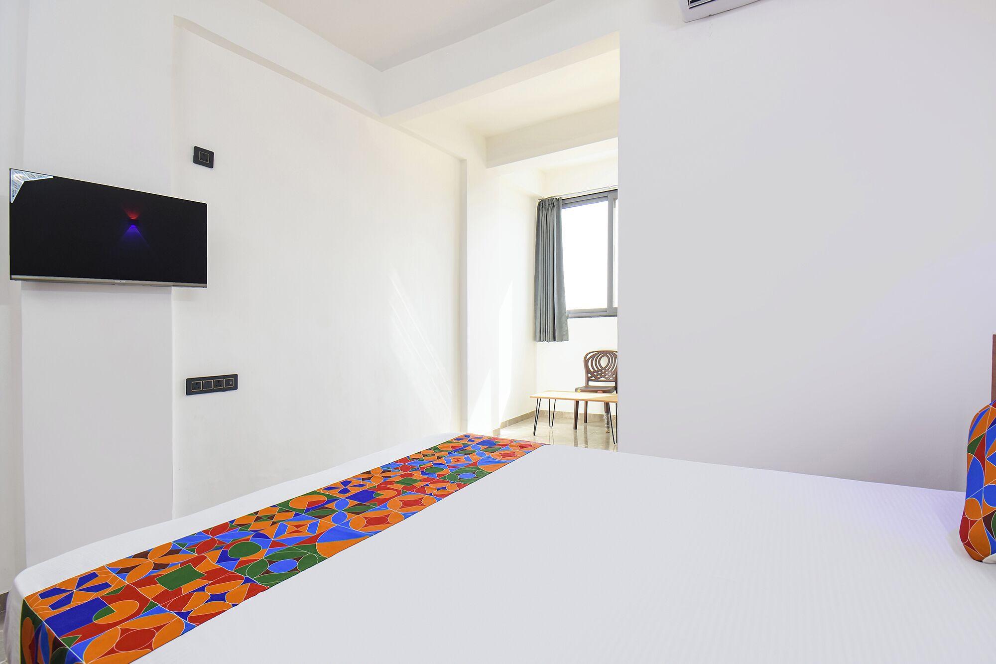 Une chambre d’hôtel avec un lit, un téléviseur fixé au mur, une fenêtre avec des rideaux et une chaise dans un coin.