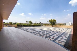 Un espacio exterior con suelo de baldosas, un árbol y un edificio blanco al fondo.