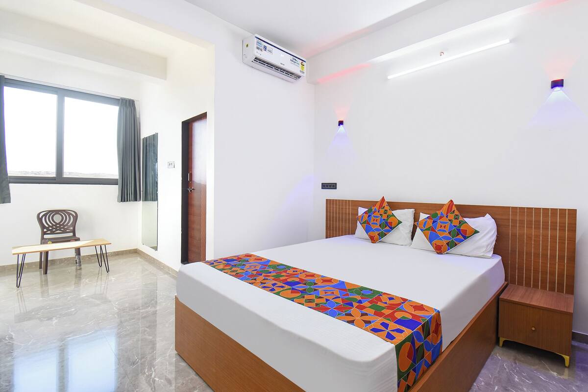 A hotel room with a bed, a bedside table, a chair, a window, and an air conditioning unit.