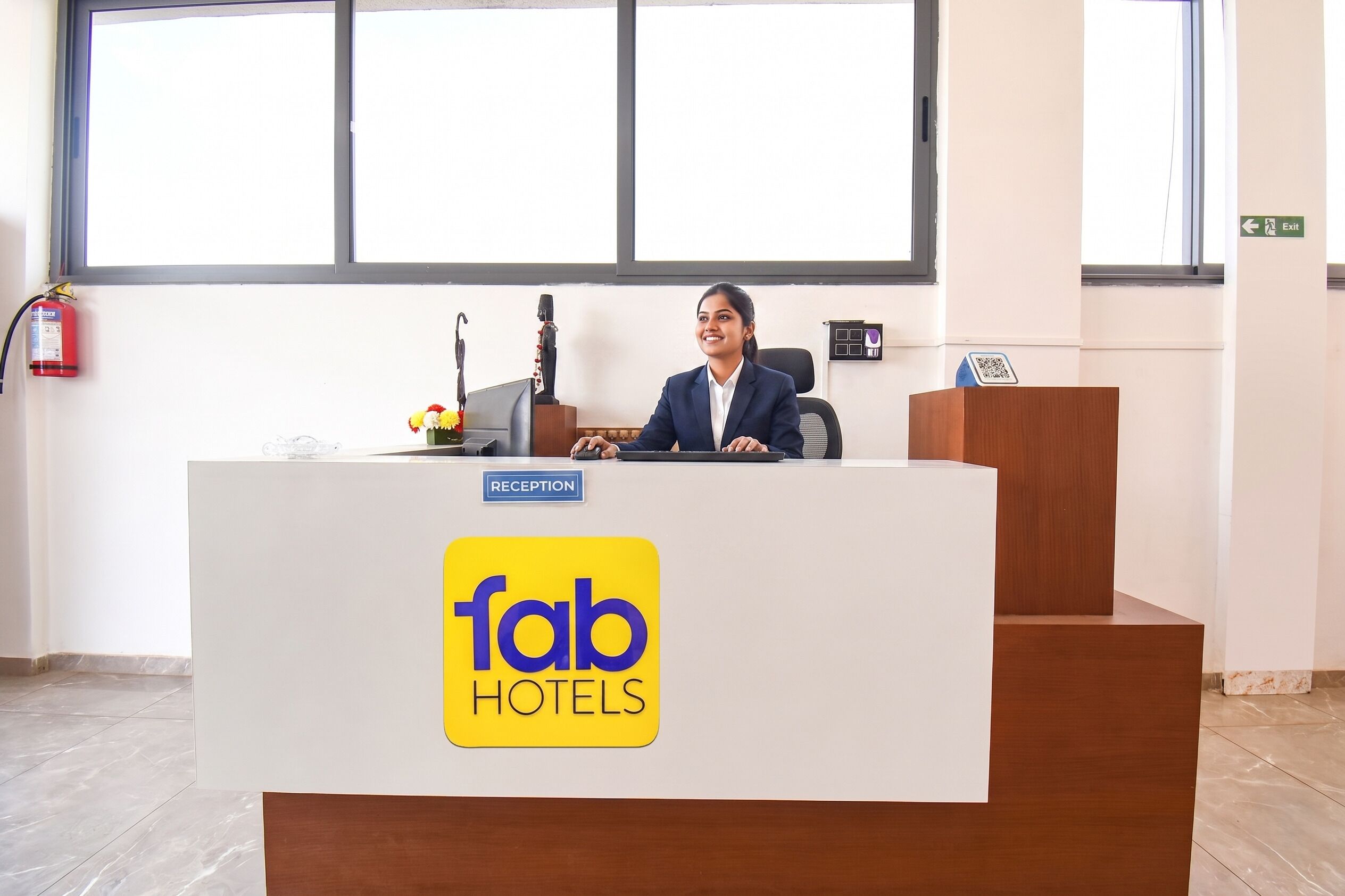 Um balcão de recepção com o logotipo 'fab hotels', uma placa com a palavra 'RECEPTION', um extintor de incêndio e uma janela com persianas.