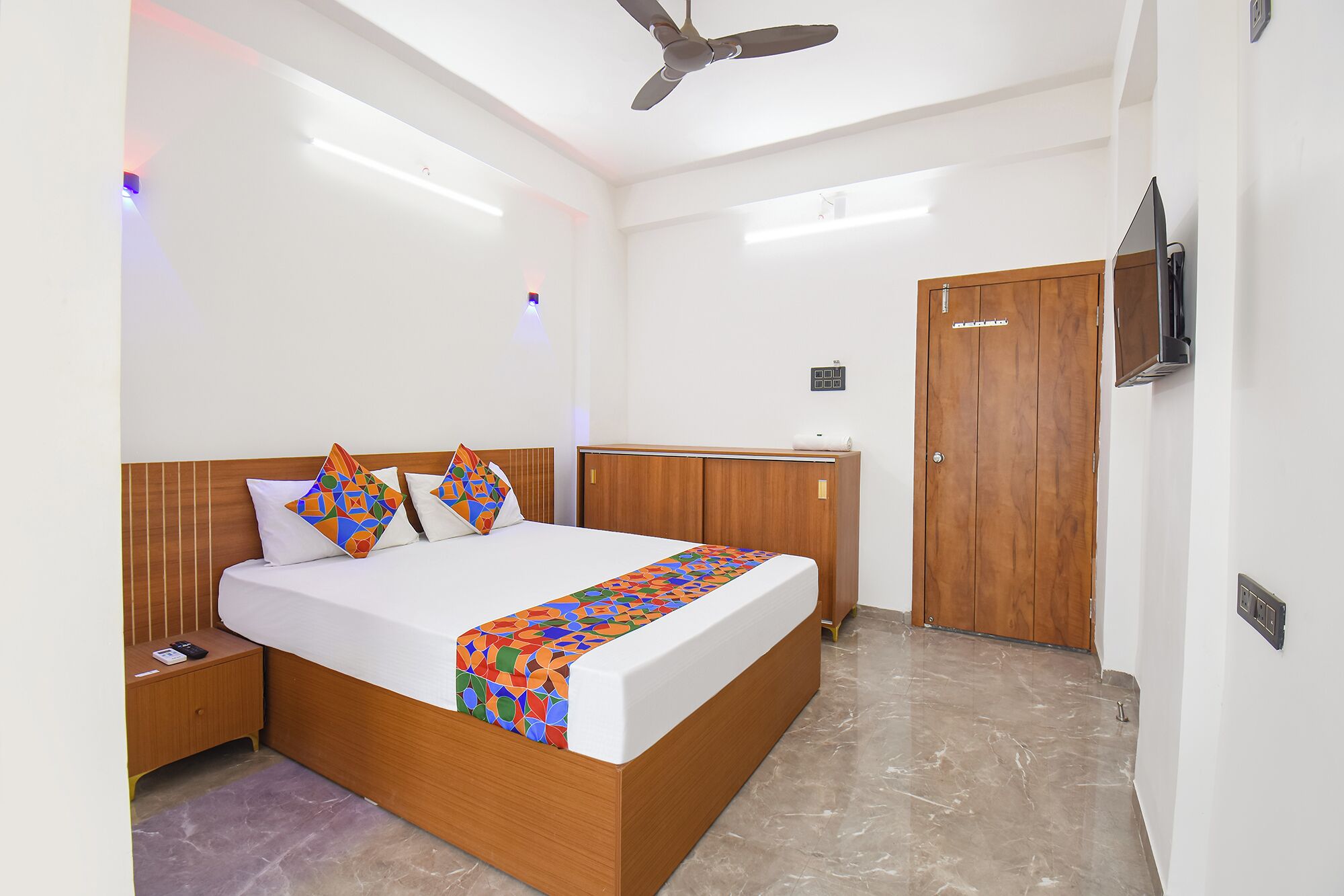 A hotel room with a bed, wooden headboard, bedside table, nightstand, TV, ceiling fan, and a door.
