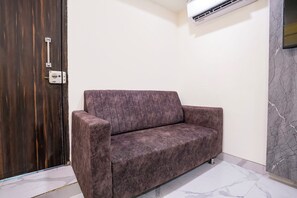 A room with a dark wooden door, a white wall, a marble floor, and a grey upholstered sofa. - Fabhotel Byland International (Mumbai)