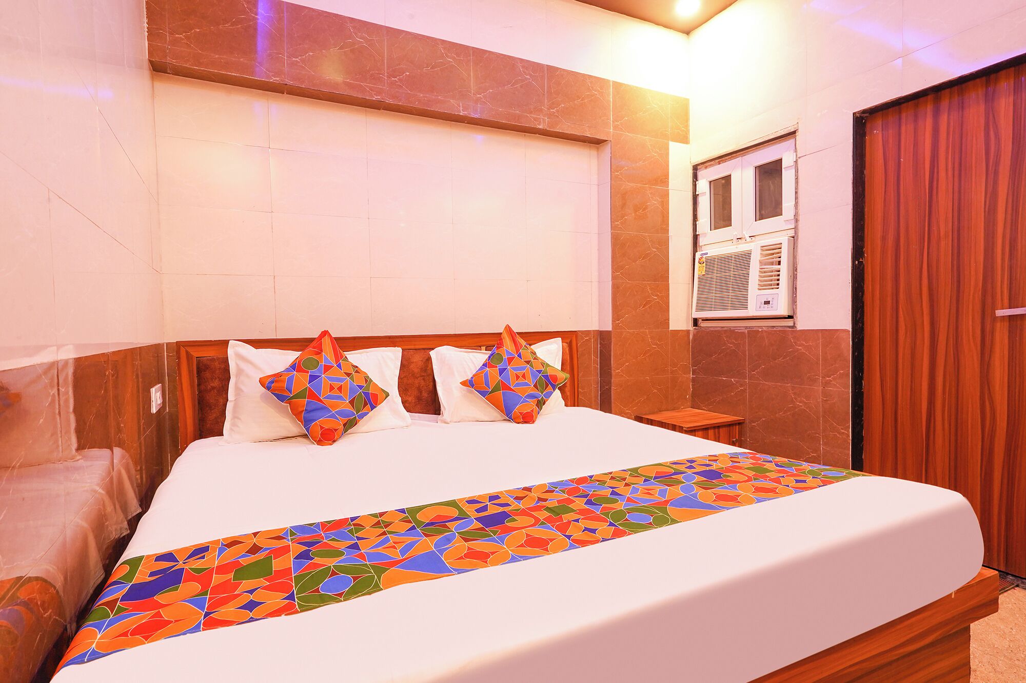 A hotel room with a bed, a wooden headboard, a window with an air conditioning unit, and a door.