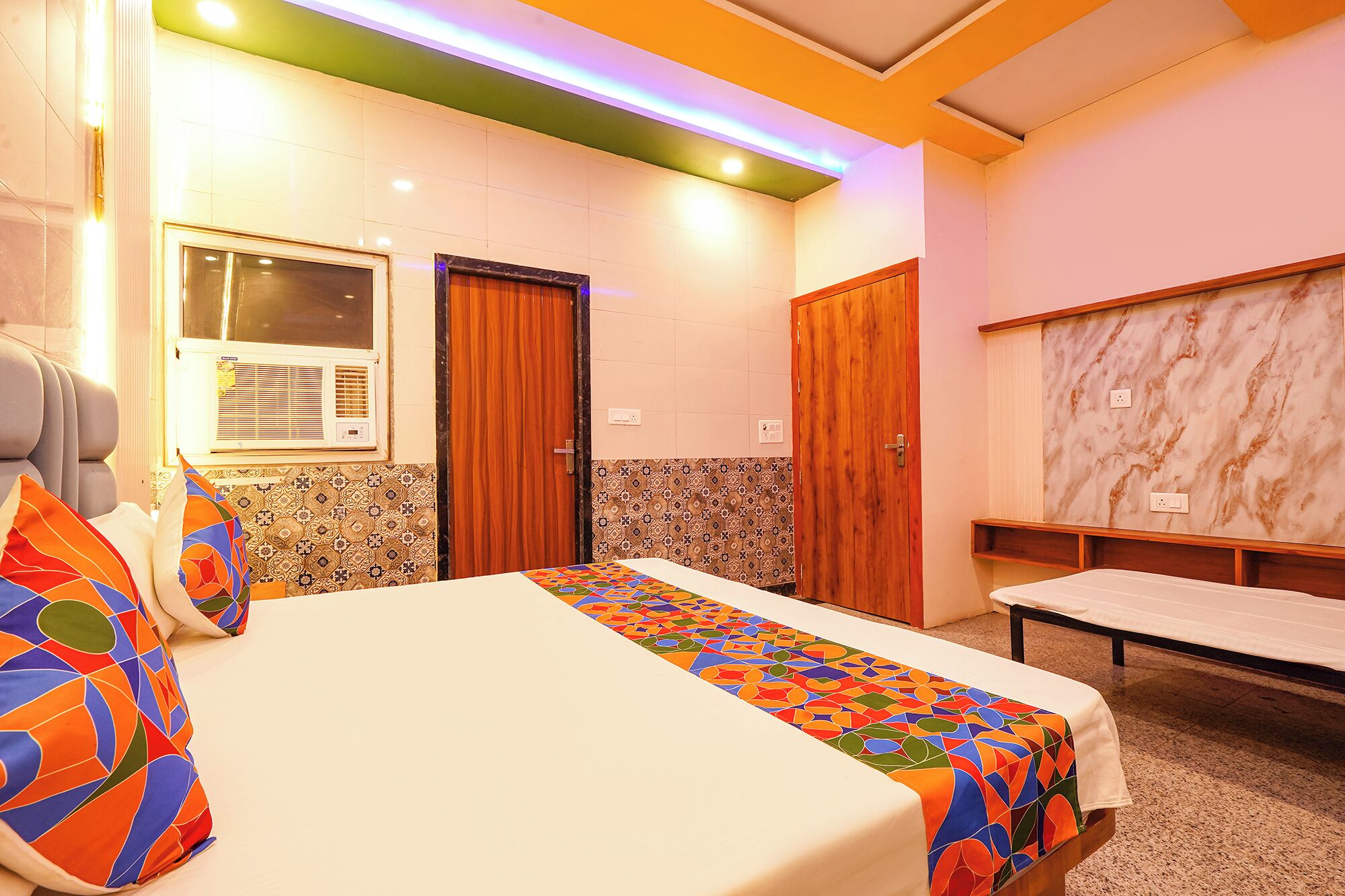 A hotel room with a bed, a window with an air conditioner, a wooden door, and a marble wall.