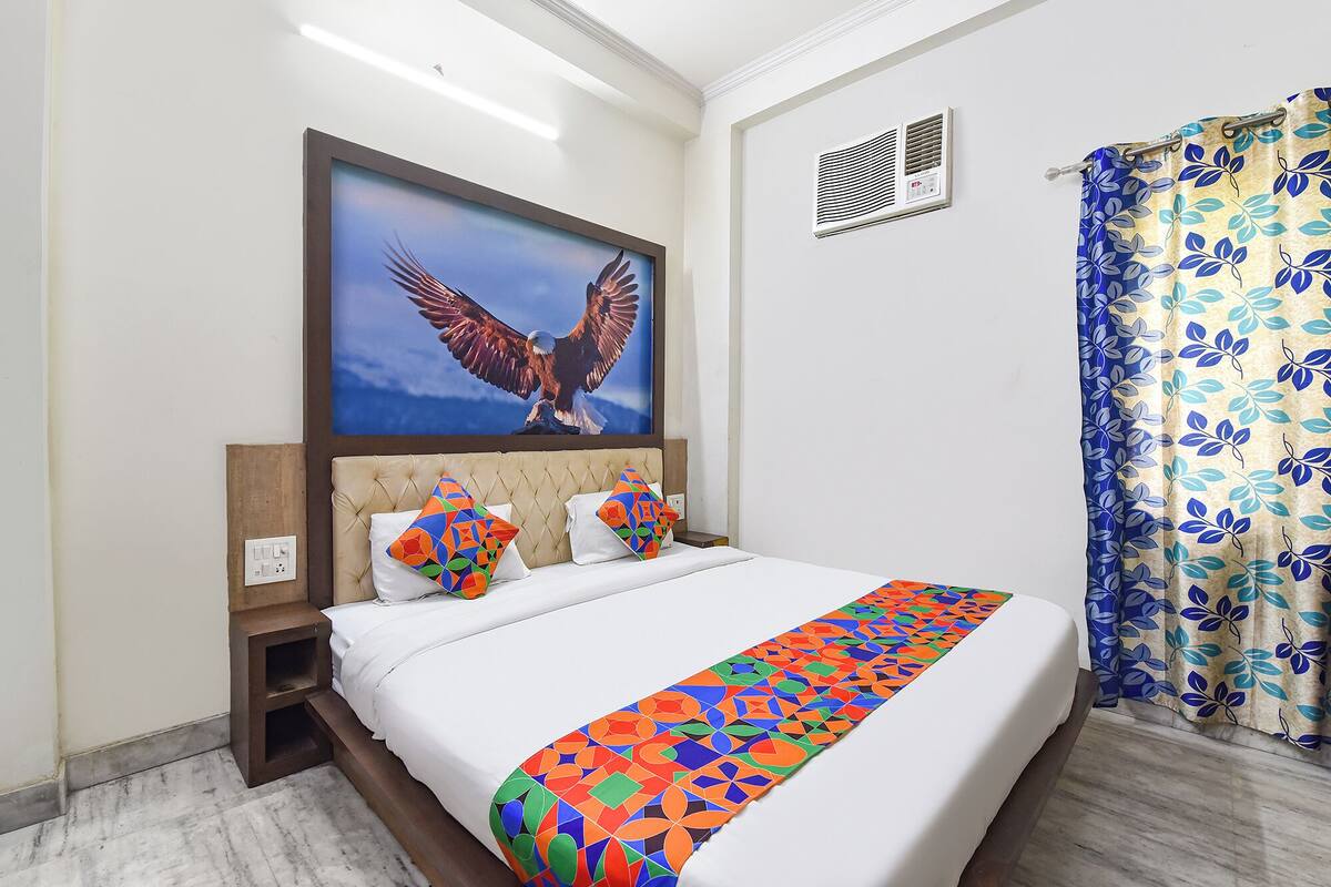 Una habitación de hotel con una cama, un cuadro grande enmarco con un águila, una ventana con cortina de estampado de hojas y un respiradero en la pared.