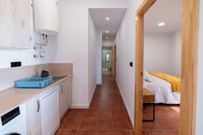 Ein schmaler Flur mit einer Küchenzeile an einer Seite, einem Bett mit gelber Decke auf der anderen und einem Badezimmer im Hintergrund.