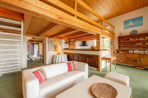 Ein gemütliches Interieur mit Holzbalkendecke, einem weißen Sofa, einem Esstisch und einer Küchenzeile mit Holzausstattung und Blick ins Freie.