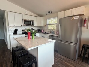 Una cocina moderna con armarios blancos, electrodomésticos de acero inoxidable, una isla central con taburetes y un extintor de incendios montado en la pared.