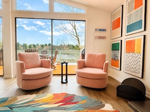 A spacious living room with two pink armchairs, a colorful rug, and large windows offering a view of trees and a deck. - "The Creel" 3BR Lakefront, Hot Tub, Resort Access (Lake Harmony)