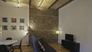 A room with a stone wall, a dining table with chairs, a sofa, a TV on a black cabinet, and framed pictures on the wall. - Apartment in Pigneto (Rome)