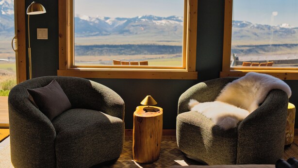 Ein Zimmer mit zwei Sesseln, einem kleinen Holztisch und einer Lampe. Ein großes Fenster bietet Blick auf Berge.