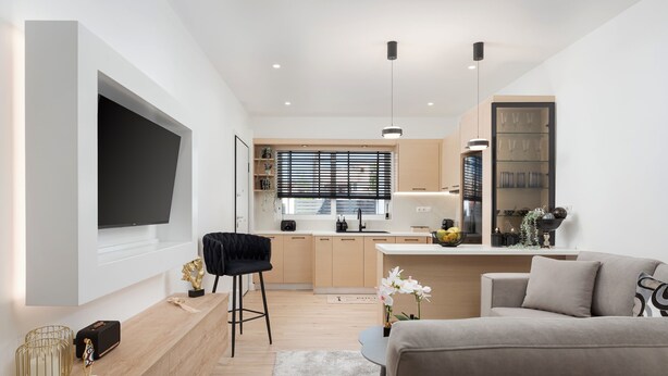 A modern living room with a large flat-screen TV, a sofa, a coffee table, and a kitchen area with a window and blinds. - Aura Airport Suite (Spata)
