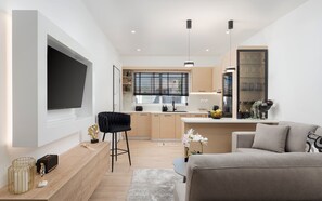 Una sala de estar moderna con un televisor de pantalla plana grande, un sofá, una mesa de centro y una zona de cocina con ventana y persianas.