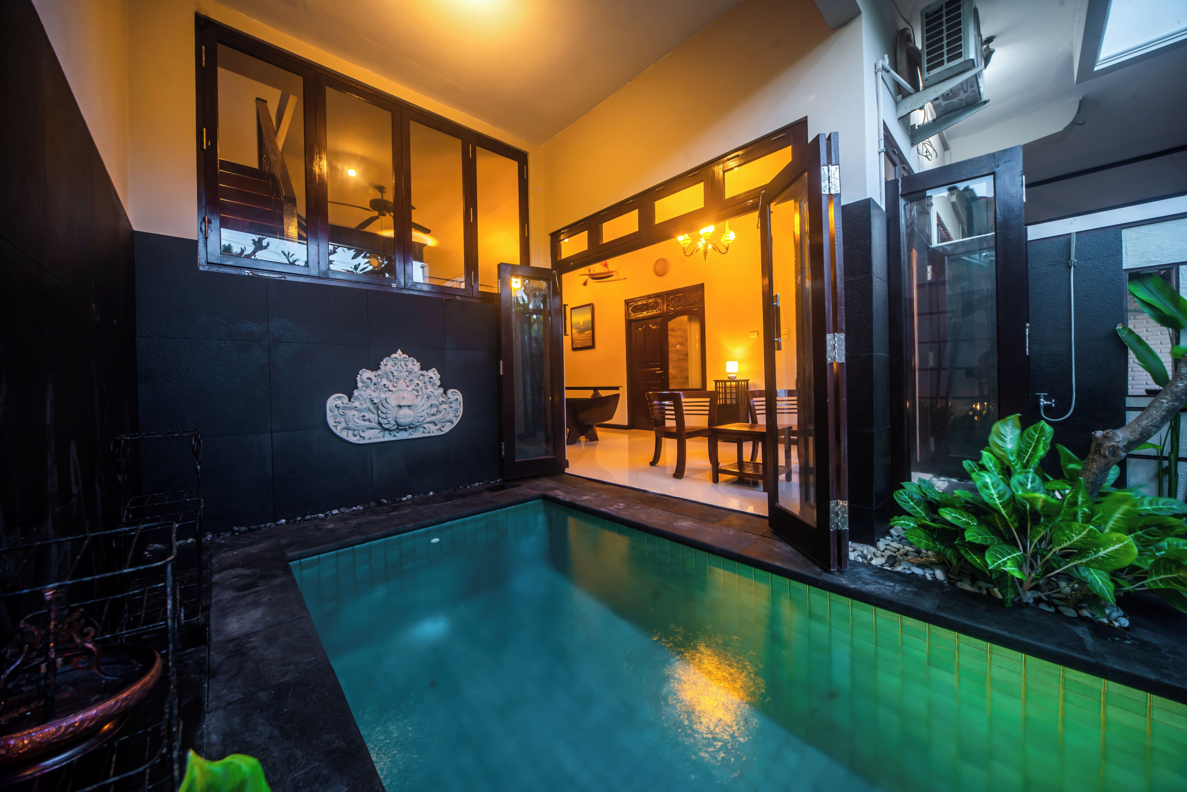 A swimming pool with a tiled edge, a dark wall with a decorative tile, and a door leading to a room with a table and chairs.