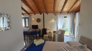 Un dormitorio con cama, un tapete azul, techo de madera, una mesa de comedor con sillas, un televisor y vistas al exterior.