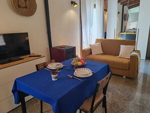 Un coin repas avec une table dressée pour deux personnes, un téléviseur posé sur un meuble, un canapé et, au fond, un espace cuisine.