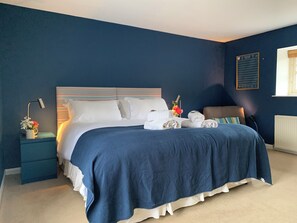 Ein ordentlich bezogenes Bett mit weißen Leinen, einem blauen Kopfteil und einem Nachttisch mit Lampe und kleiner Blumenvase.