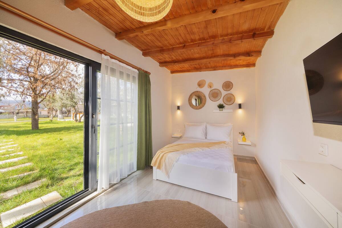 Une chambre avec une grande fenêtre, un lit, une table de chevet et un téléviseur fixé au mur.