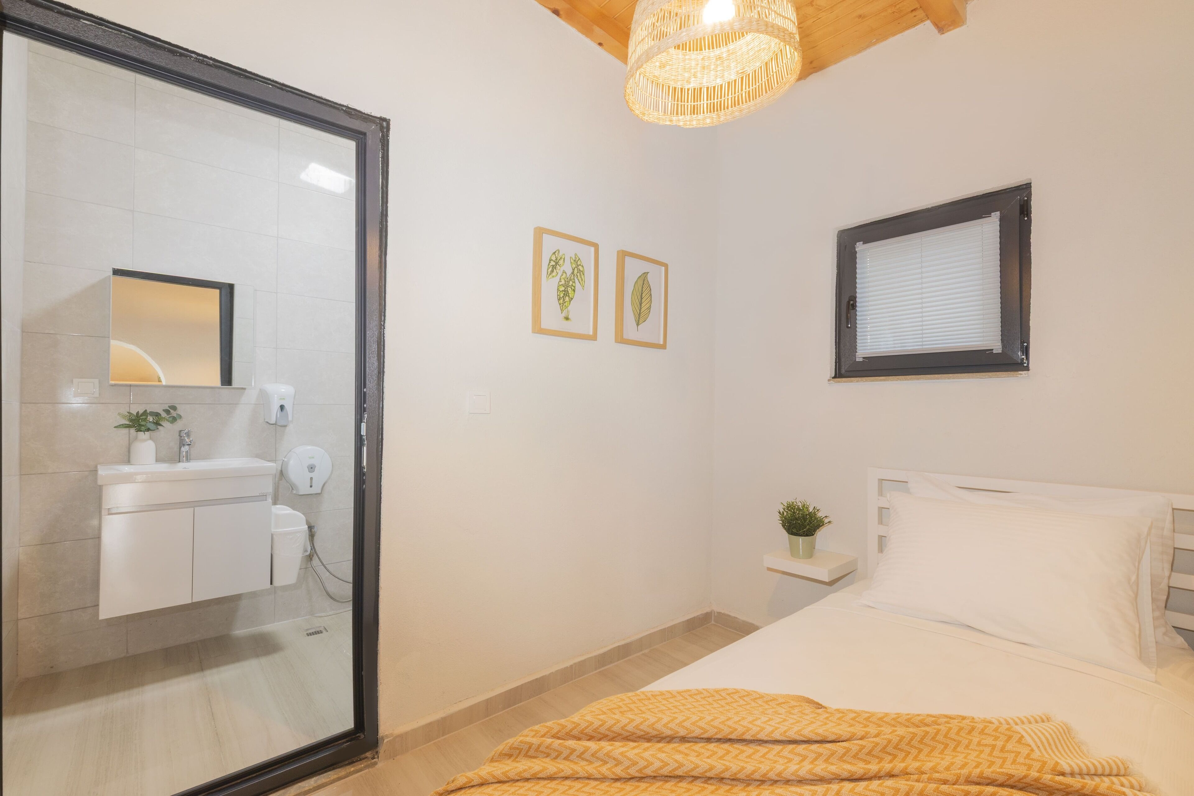 A bedroom with a bed, a bedside table with a plant, a window with blinds, and a bathroom with a mirror, sink, and towel rack.
