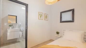 A bedroom with a bed, a bedside table with a plant, a window with blinds, and a bathroom with a mirror, sink, and towel rack.