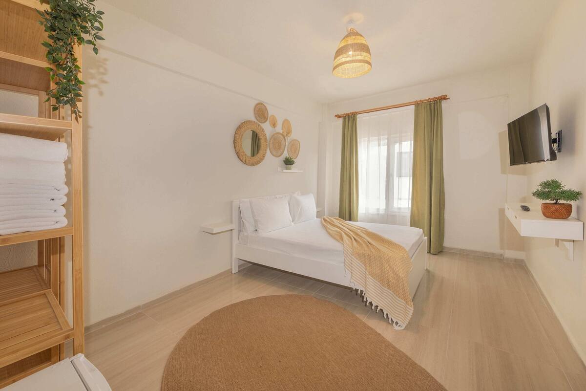 Une chambre d’hôtel avec un lit, une télévision, une étagère avec des serviettes, une plante suspendue et une grande fenêtre avec des rideaux.