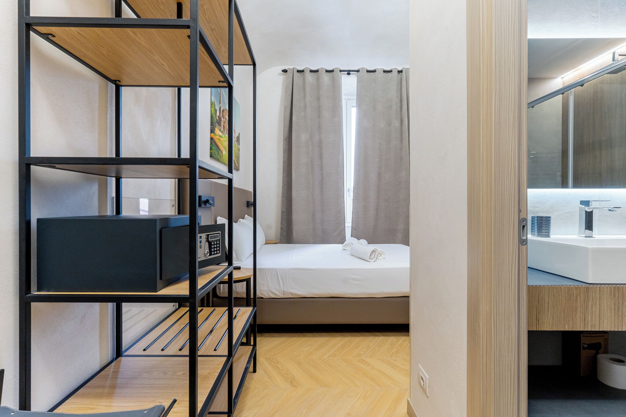 A compact hotel room with a bunk bed, a desk with a computer, a bedside table, a window with curtains, a bathroom with a mirror and sink, and a wooden floor.