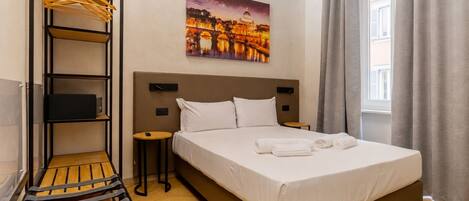 A hotel room with a bed, a wooden bench, a shelf with hangers, a small table, and a painting of a bridge.