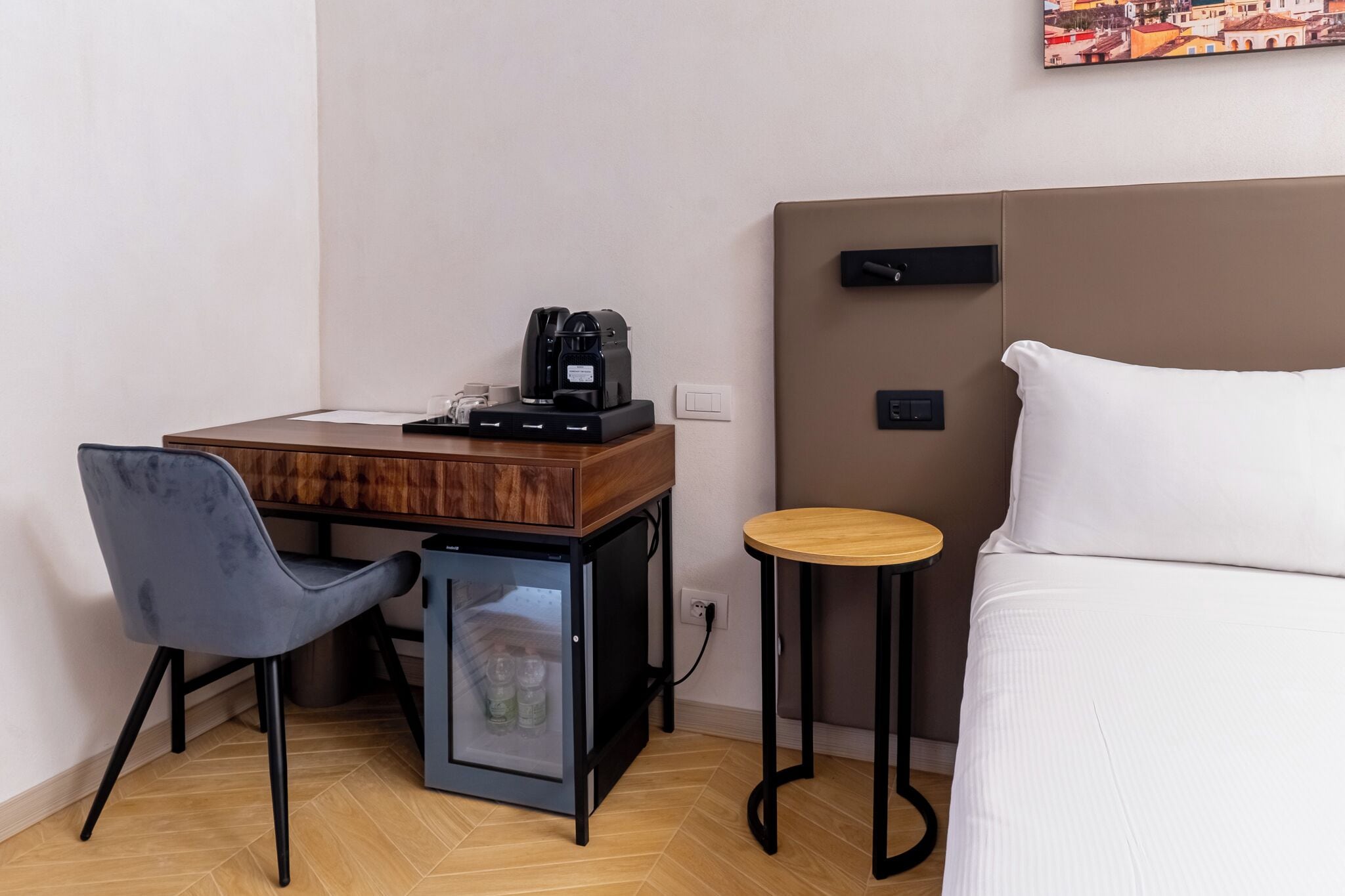 A compact hotel room with a bed, a small desk with a coffee maker, a chair, and a mini-fridge.