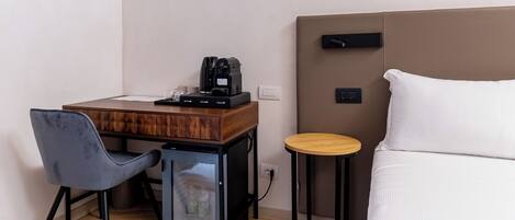 A compact hotel room with a bed, a small desk with a coffee maker, a chair, and a mini-fridge.