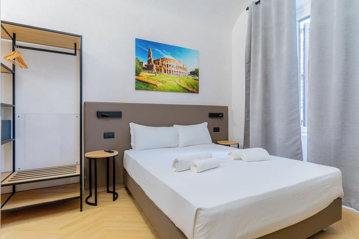 Hotellihuone, jossa on sänky, pieni pöytä, vaatteita varten tarkoitettu hylly, rakennuksen kuva ja ikkuna, jossa on verhot.