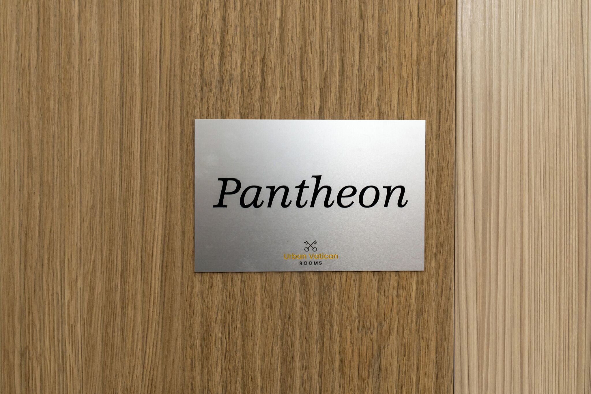 A metallic sign with the word 'Pantheon' and the logo 'Urban Vatican ROOMS' on a wooden surface.