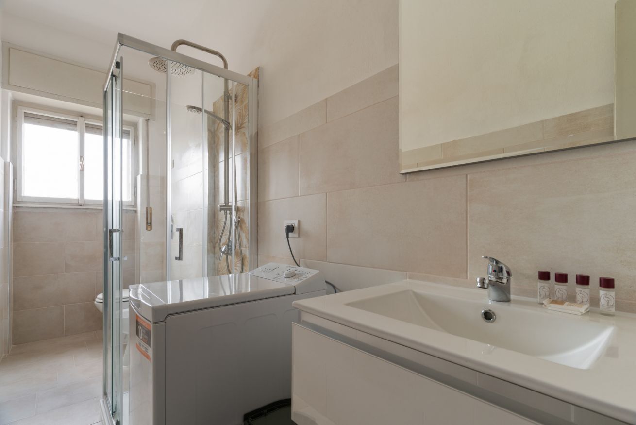 A modern bathroom with a glass shower enclosure, a built-in washing machine, a large sink, and a mirror above the sink.