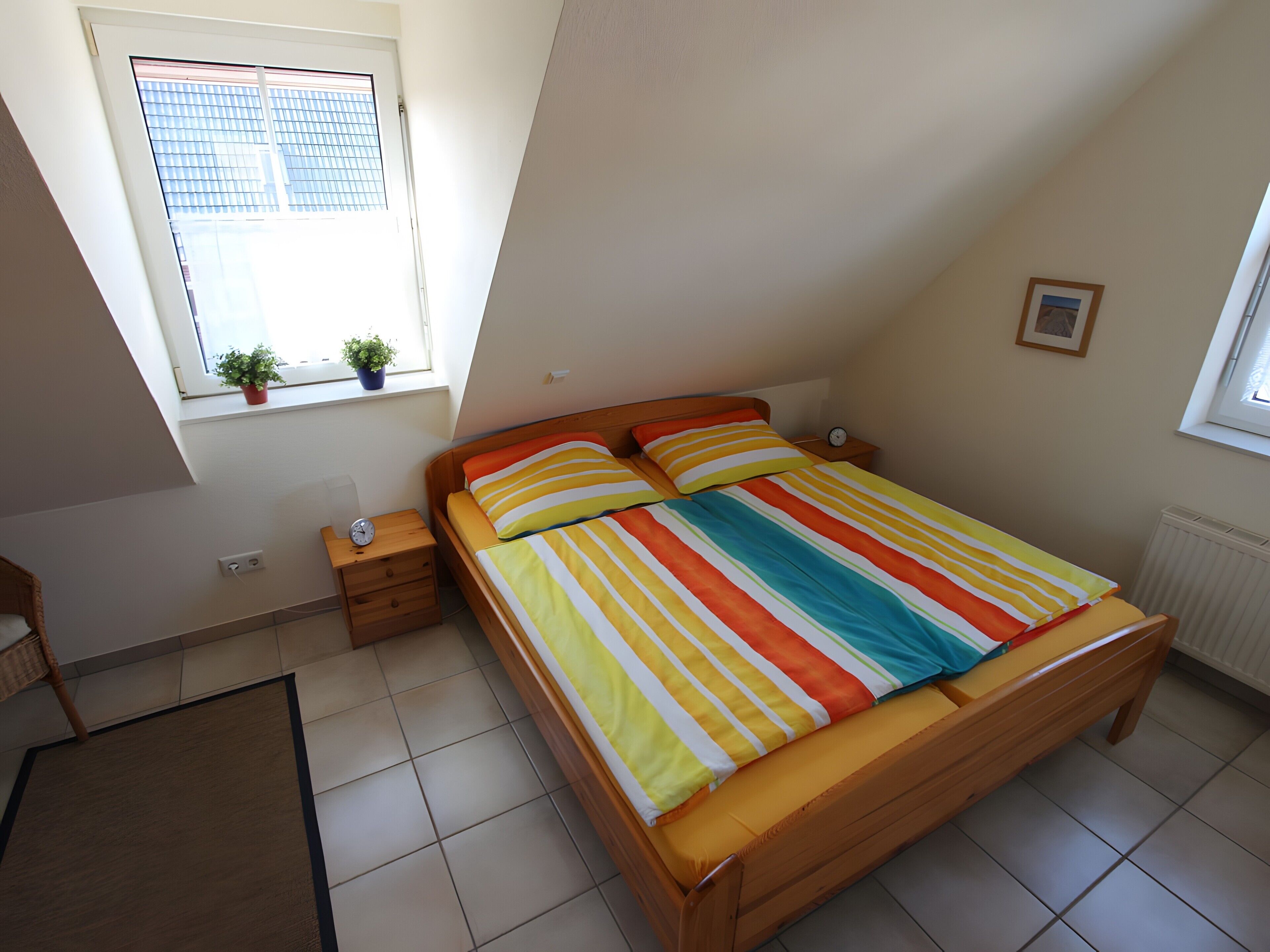 Ein Schlafzimmer mit geneigter Decke, einem Bett mit gestreifter Bettwäsche, einem Nachttisch mit einer Uhr, einer kleinen Topfpflanze und einem Fenster mit Jalousien.