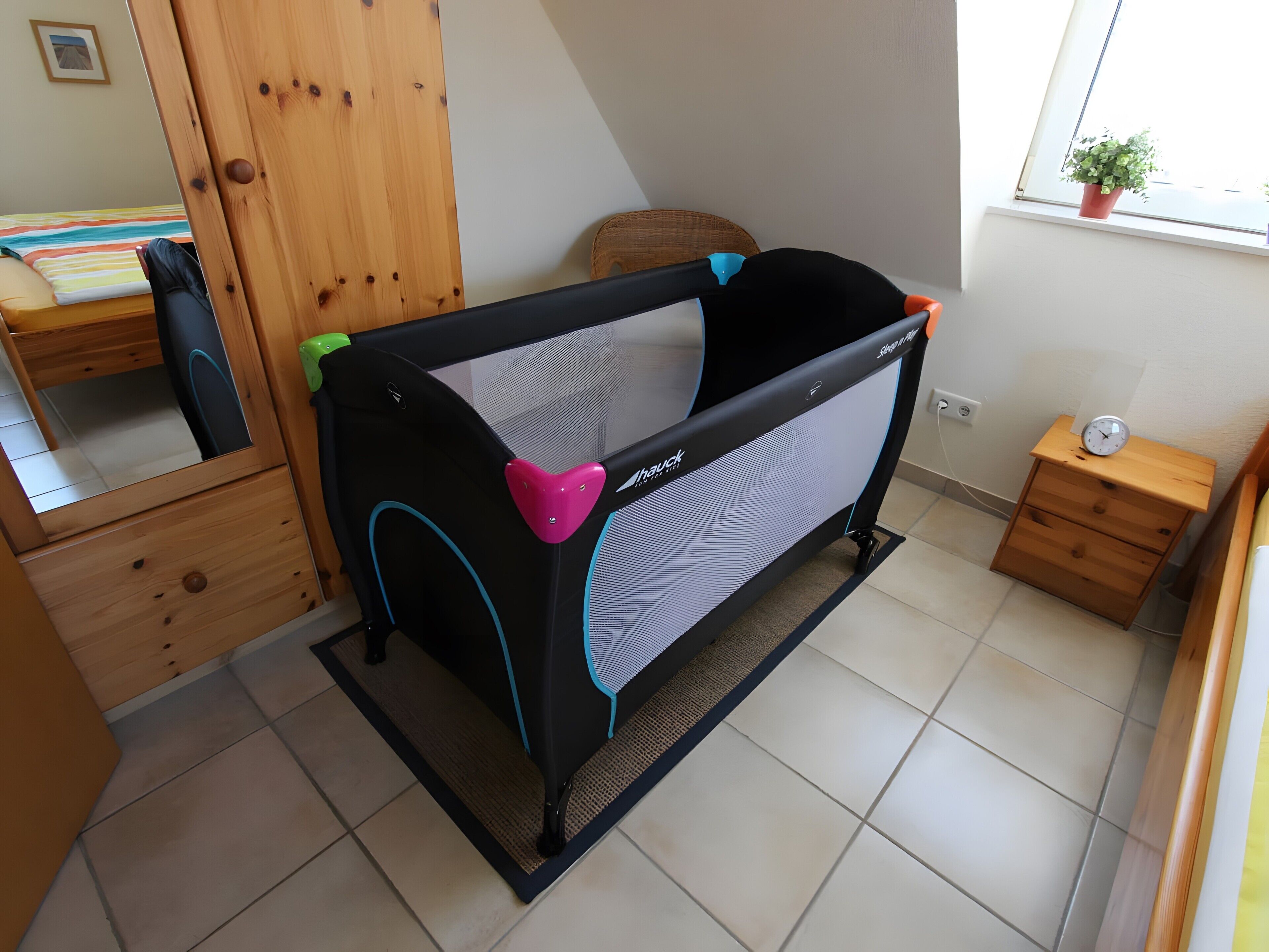 Une pièce avec un parc pour bébé noir et gris, une armoire en bois, une petite table de chevet en bois et une fenêtre avec une plante.
