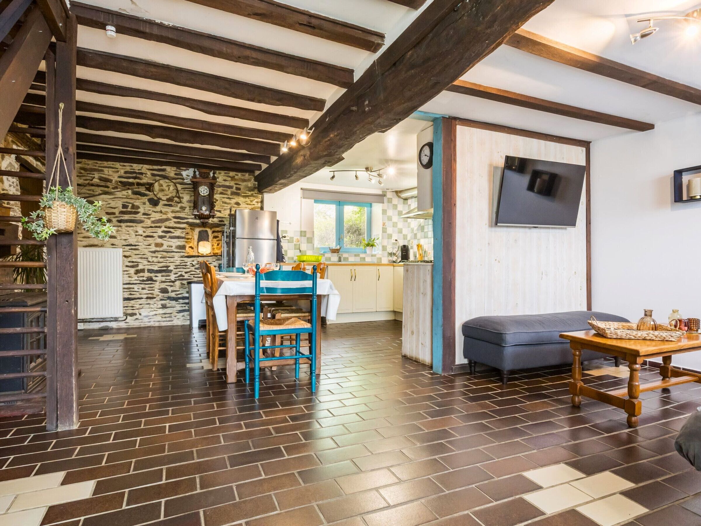 Un intérieur rustique avec un mur en pierre, des poutres en bois, un coin repas avec une table et des chaises, une cuisine à l’arrière-plan et un canapé au premier plan.
