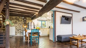 Un interior rústico con pared de piedra, vigas de madera, una zona de comedor con mesa y sillas, una cocina al fondo y un sofá en primer plano.