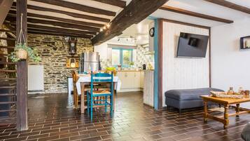 Un intérieur rustique avec un mur en pierre, des poutres en bois, un coin repas avec une table et des chaises, une cuisine à l’arrière-plan et un canapé au premier plan.
