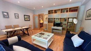 Una sala de estar con sofá, mesa de centro, televisor, estantería, mesa de comedor, sillas y cuadros en la pared.